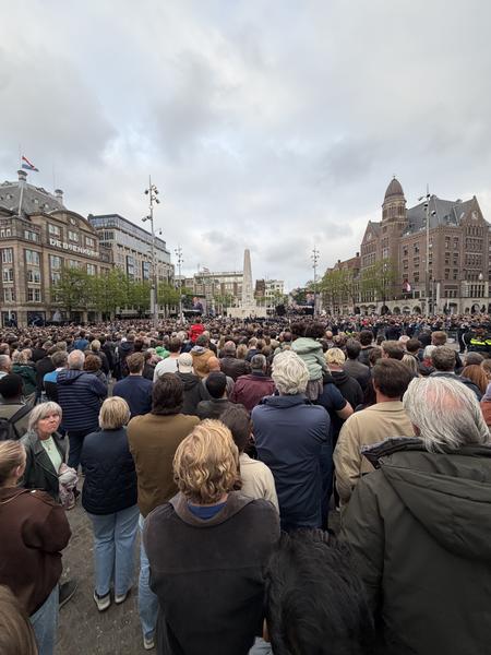 A vast crowd fills Dam Square in Amsterdam, facing towards the National Monument under an overcast sky